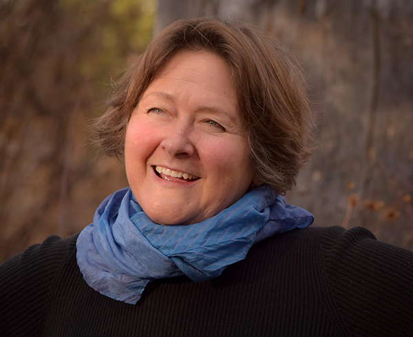 A smiling Susan Strauss with short brown hair, a blue scarf and black shirt and a green and brown winter landscape behind her.